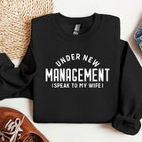 a black sweater that says under new management i speak to my wife